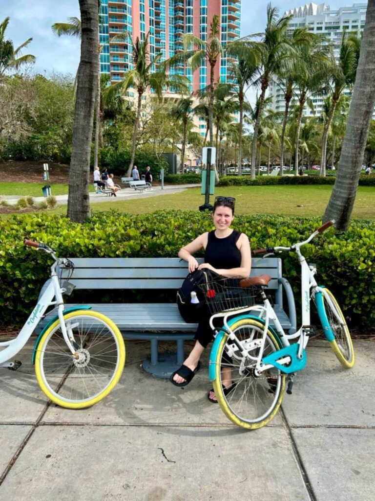 Alice Hobbs sits on a bench in a park next to two bicycles under palm trees in Miami